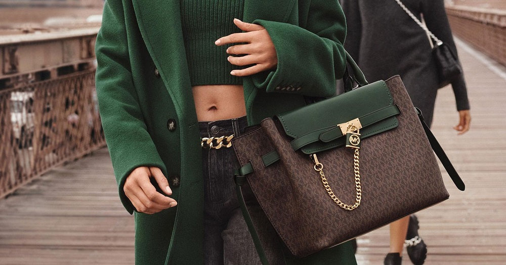 Women carrying Michael Kors sales bags green and brown with gold colour chain and lock. Walking across wooden bridge.